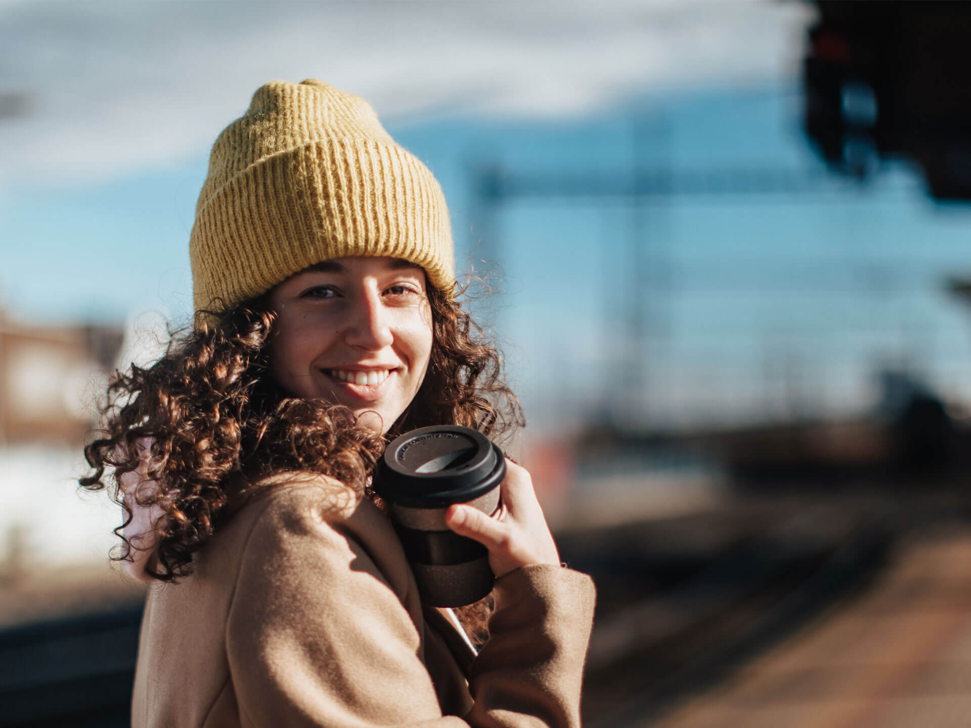 Junge Frau steht am Bahngleis und hält einen Kaffeebehälter in der Hand.