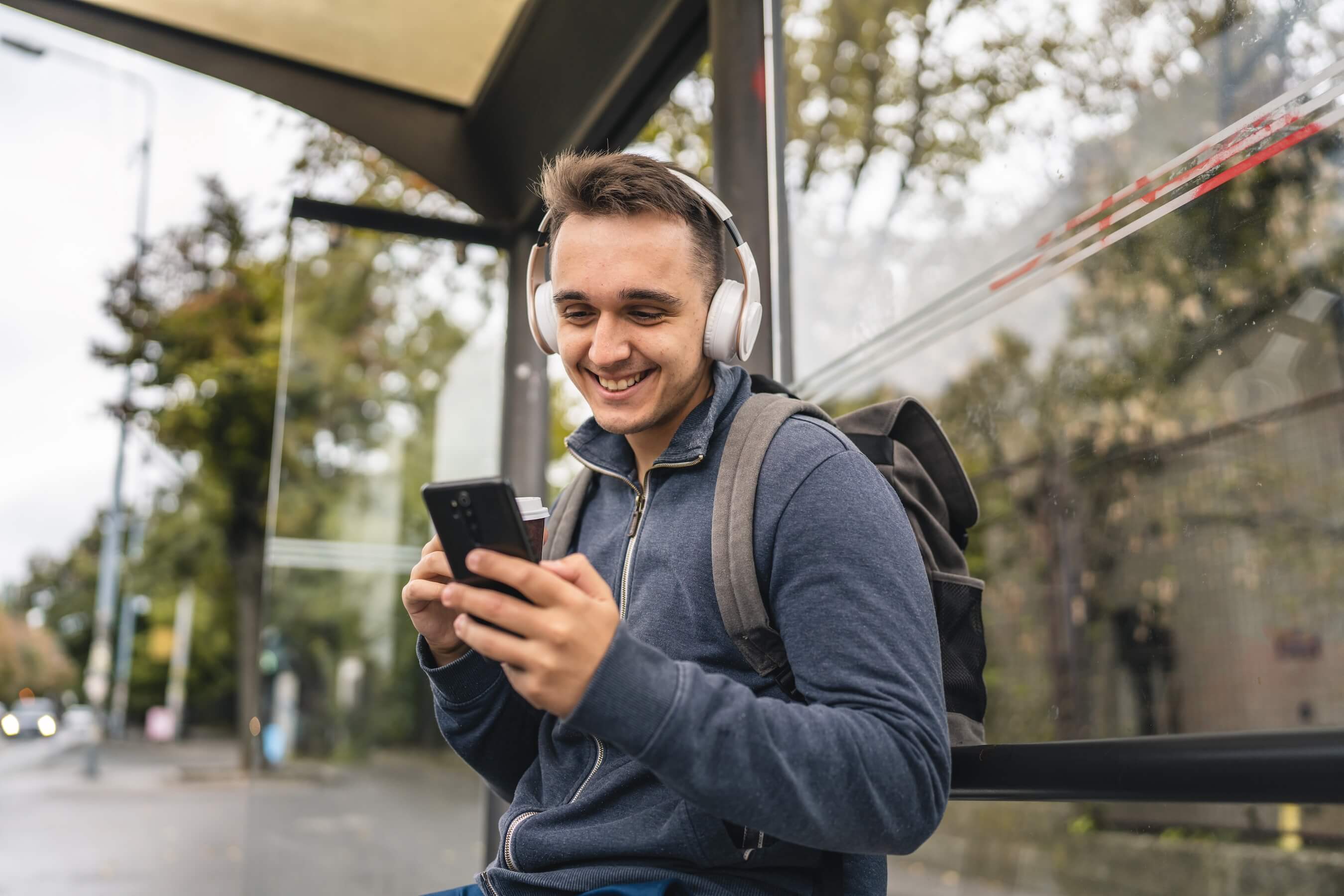 Ein junger Mann der Kopfhörer trägt, steht an einer Bushaltestelle und schaut auf sein Smartphone.