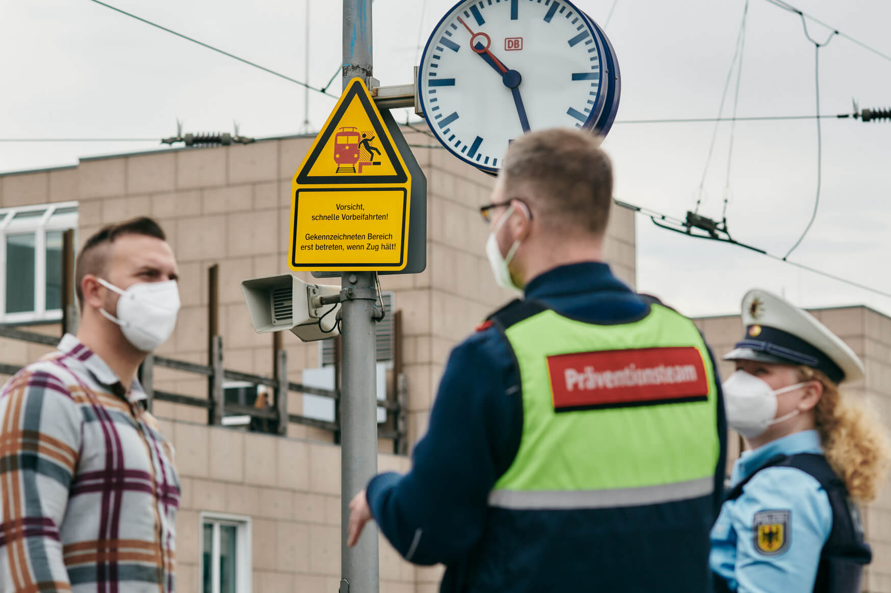 Ein DB-Mitarbeiter in einer gelben Weste, eine Polizei-Beamtin und ein junger Mann stehen unter einer Uhr auf einem Bahnsteig.