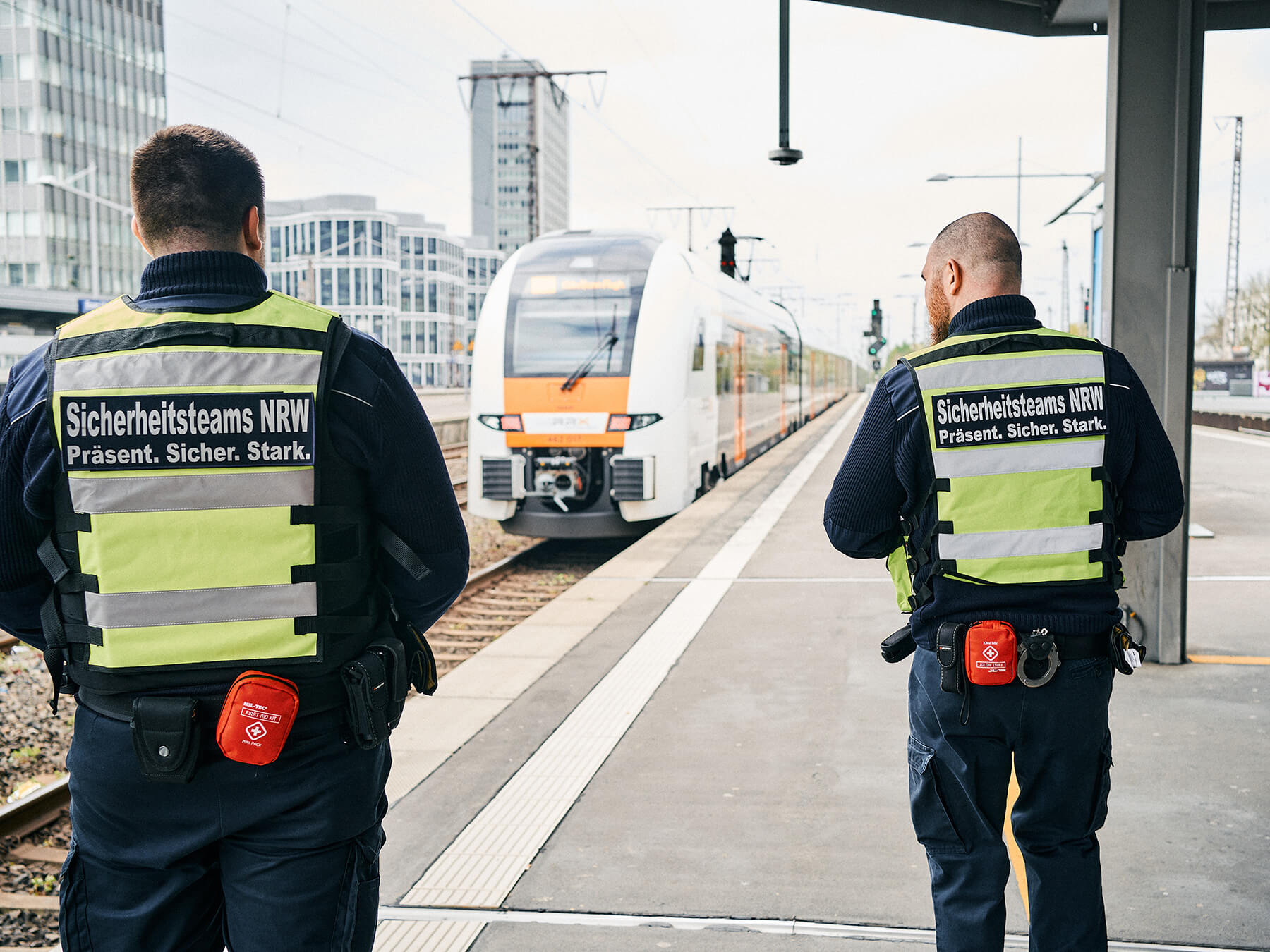 Zwei Männer in gelben Sicherheitswesten laufen an einem Bahnsteig entlang, an dem ein Zug steht.