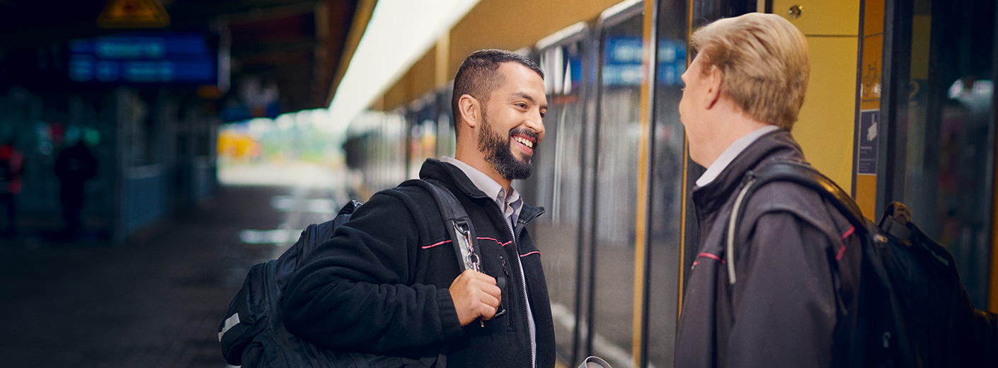 Zwei Lokführer unterhalten sich und stehen an einem Bahnsteig, an dem ein Zug hält. 