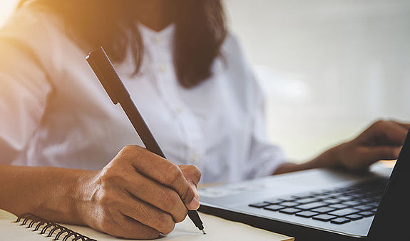 Eine Frau sitzt an einem Schreibtisch. Vor ihr steht ein geöffneter Laptop und sie schreibt mit einem Kugelschreiber auf einen Block.