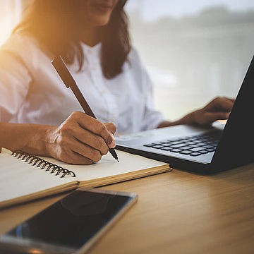 Eine Frau sitzt an einem Schreibtisch. Vor ihr steht ein geöffneter Laptop und sie schreibt mit einem Kugelschreiber auf einen Block.
