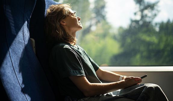 Eine Frau mit Brille sitzt in mit geschlossenen Augen in einem Zug während am Fenster eine grüne Landschaft vorbeizieht.