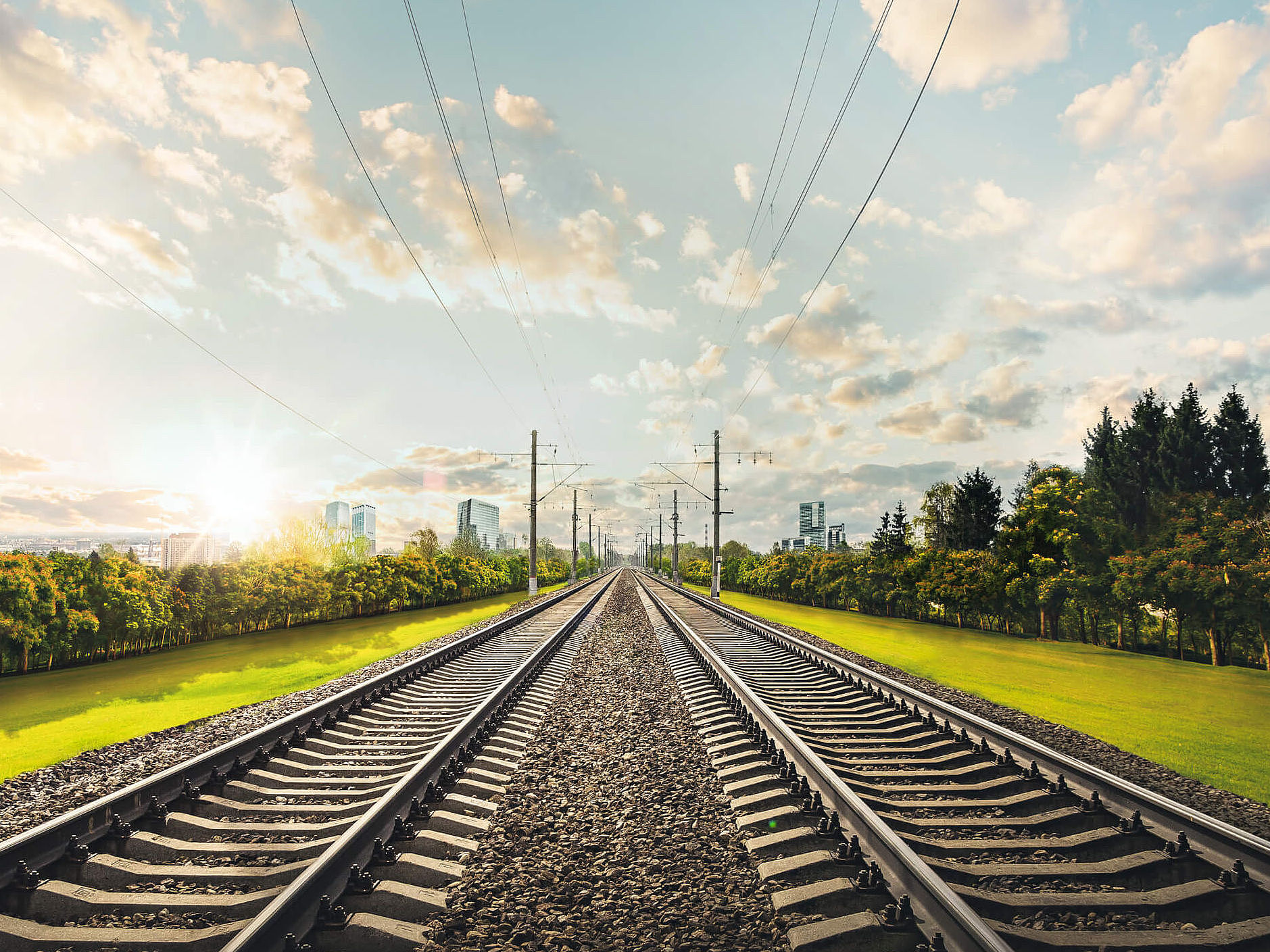 Zwei Schienen verschwinden im Horizont, links und rechts ist grünes Gras zu sehen und die Sonne scheint. Am Horizont sind moderne Gebäuder erkennbar.