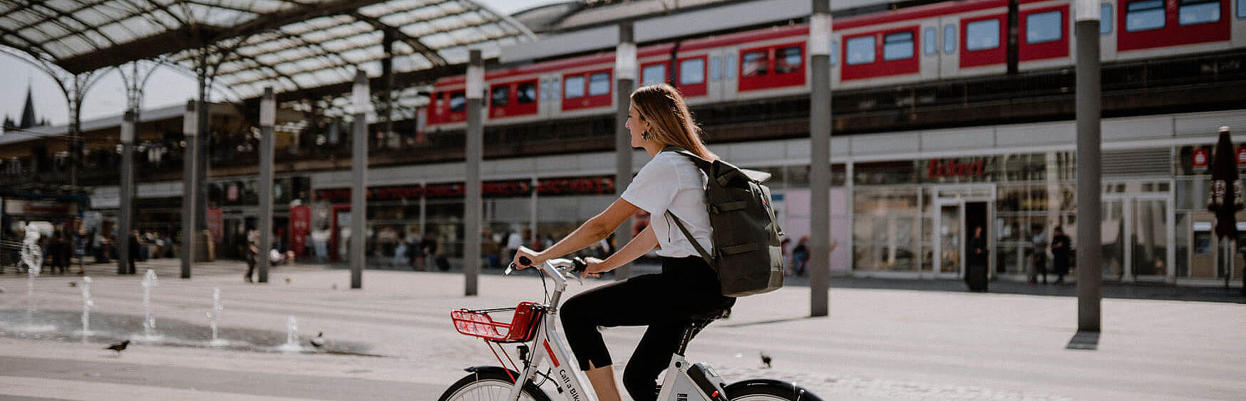 Eine Frau auf einem Mietrad fährt an einem Bahnhof vorbei. Im Hintergrund sieht man einen roten Zug auf einer Eisenbahnbrücke.