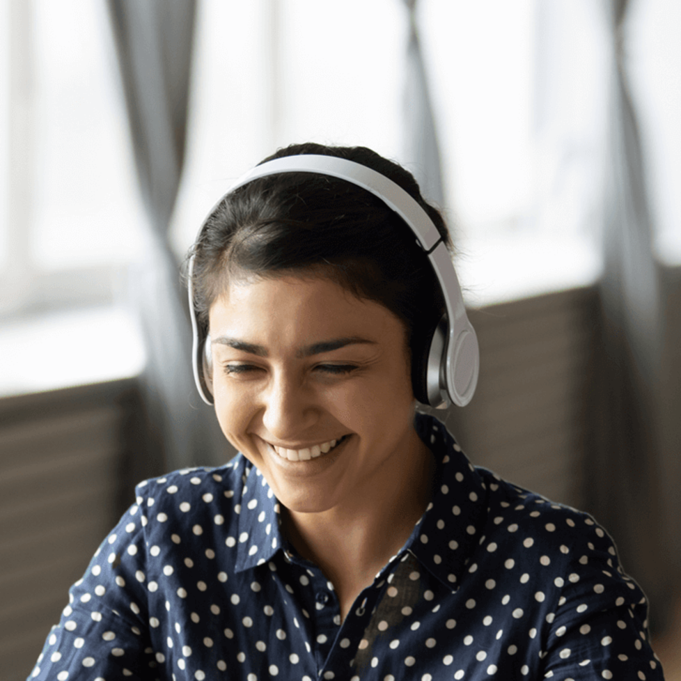 Eine junge Frau sitzt mit einem Headset vor einem Laptop in ihrem Wohnzimmer und führt ein Gespräch per Videocam.