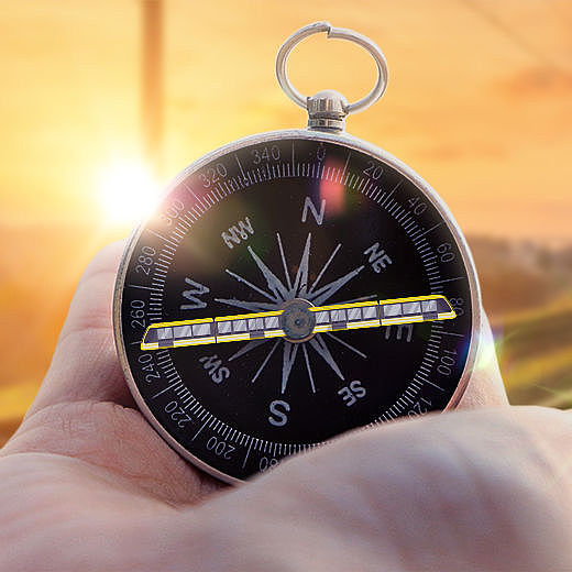 Eine Hand hält einen Kompass auf dem ein Zug abgebildet ist. Im Hintergrund sind Bahnschienen bei Sonnenuntergang zu sehen.