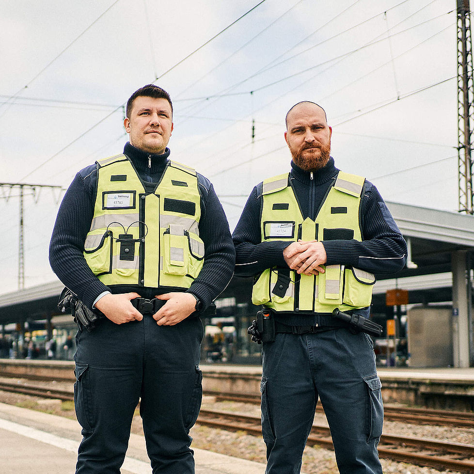 Zwei Männer in gelben Sicherheitswesten stehen an einem Bahnsteig und schauen in die Kamera.