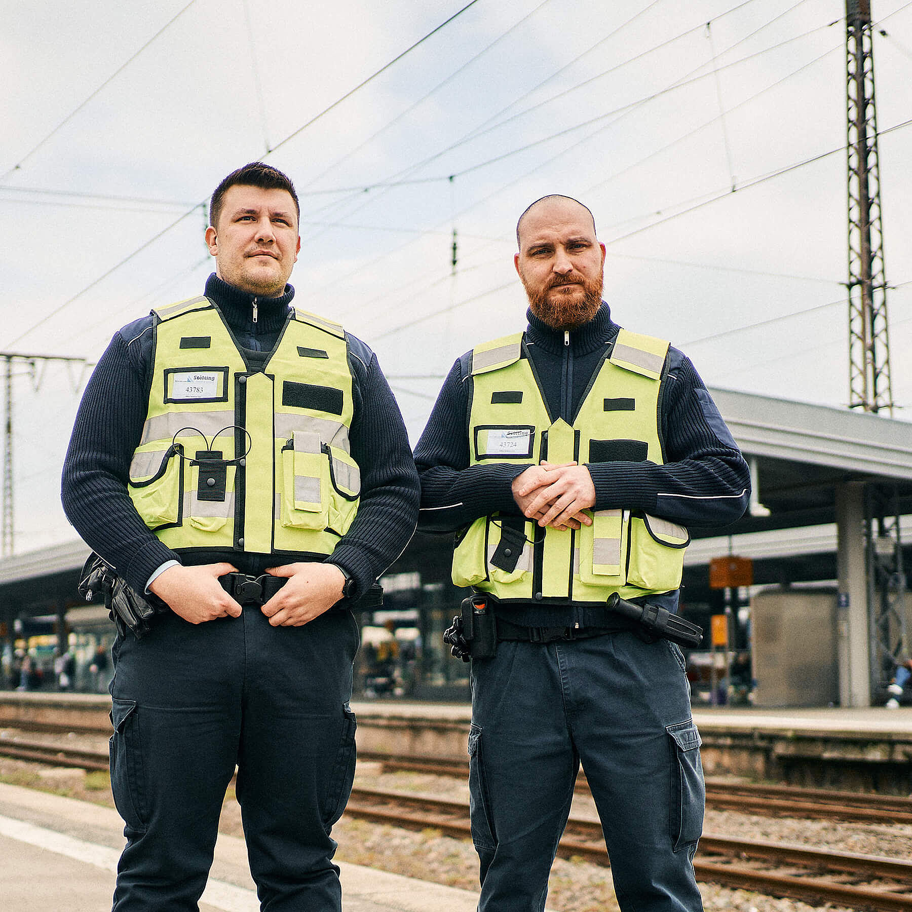 Zwei Männer in gelben Sicherheitswesten stehen an einem Bahnsteig und schauen in die Kamera.