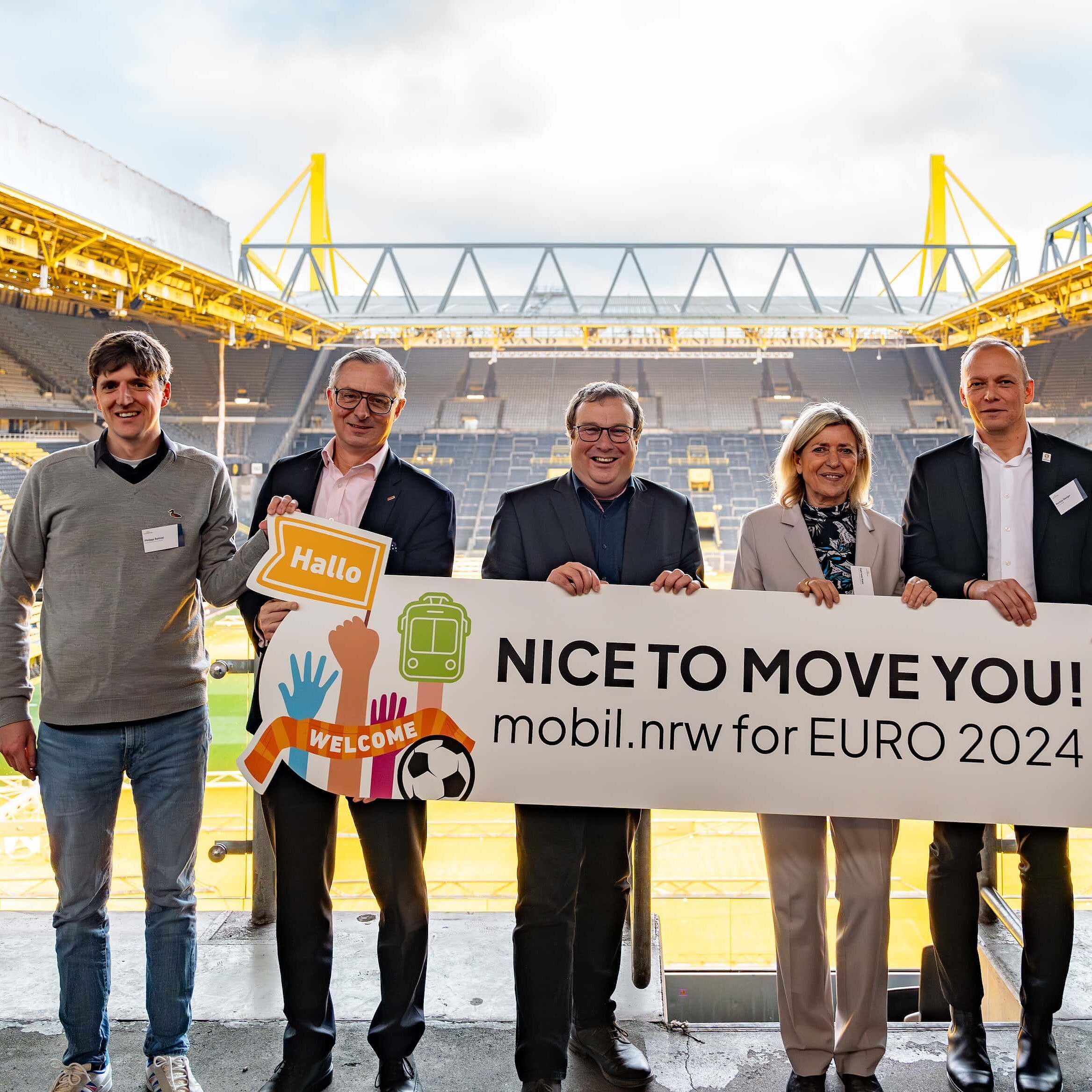 Mehrere Personen stehen auf der Tribüne eines Stadions und halten ein Schild mit der Aufschrift "Nice to move you!" hoch – dem Motto des NRW-Nahverkehrs zur EURO 2024.