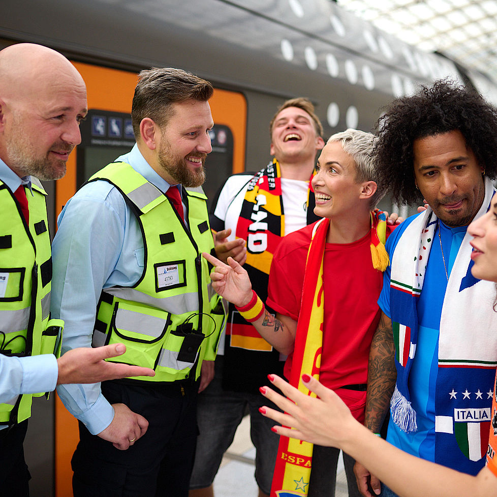 Internationale Fußballfans in Fankleidung stehen an einem Bahnsteig und sprechen mit Mitarbeitern des Sicherheitsteams NRW.