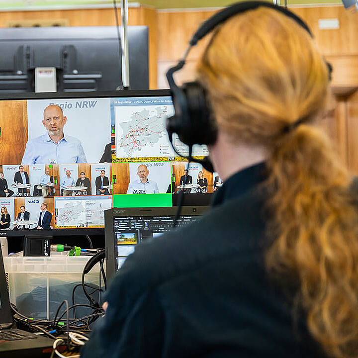Ein Mann mit roten langen Haaren und einem Headset sitzt an mehreren Monitoren und überwacht die Aufnahme des Live-Streams.