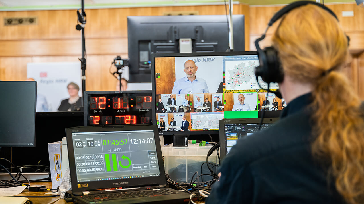 Ein Mann mit roten langen Haaren und einem Headset sitzt an mehreren Monitoren und überwacht die Aufnahme des Live-Streams.