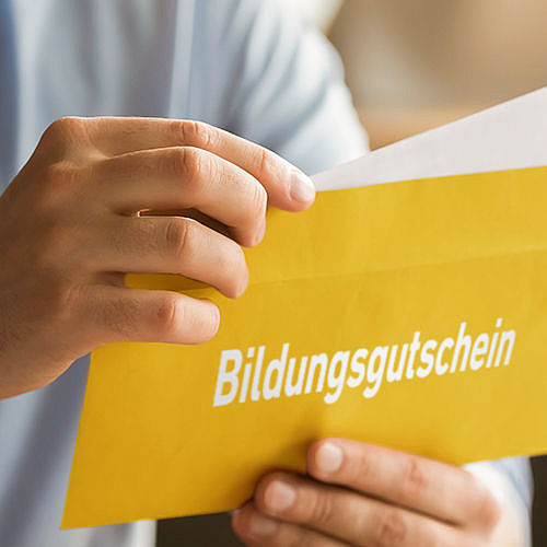 Ein Mann hält einen gelben Umschlang in der Hand und öffnet ihn. Auf dem Umschlag ist in weißen Buchstaben das Wort "Bildungsgutschein" zu lesen.