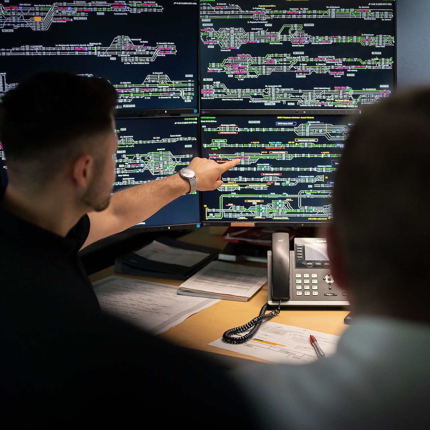 Zwei Männer sitzen vor einem Schreibtisch auf dem mehrere, große Monitore mit Daten stehen. 