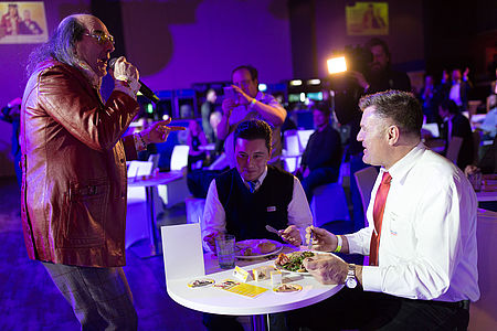 Guildo Horn steht singend vor zwei Männern in weißen Hemden, die ihm zuhören und dabei an einem Tisch etwas essen.