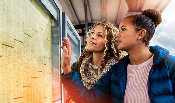 Zwei junge Frauen informieren sich an einem Fahrplan