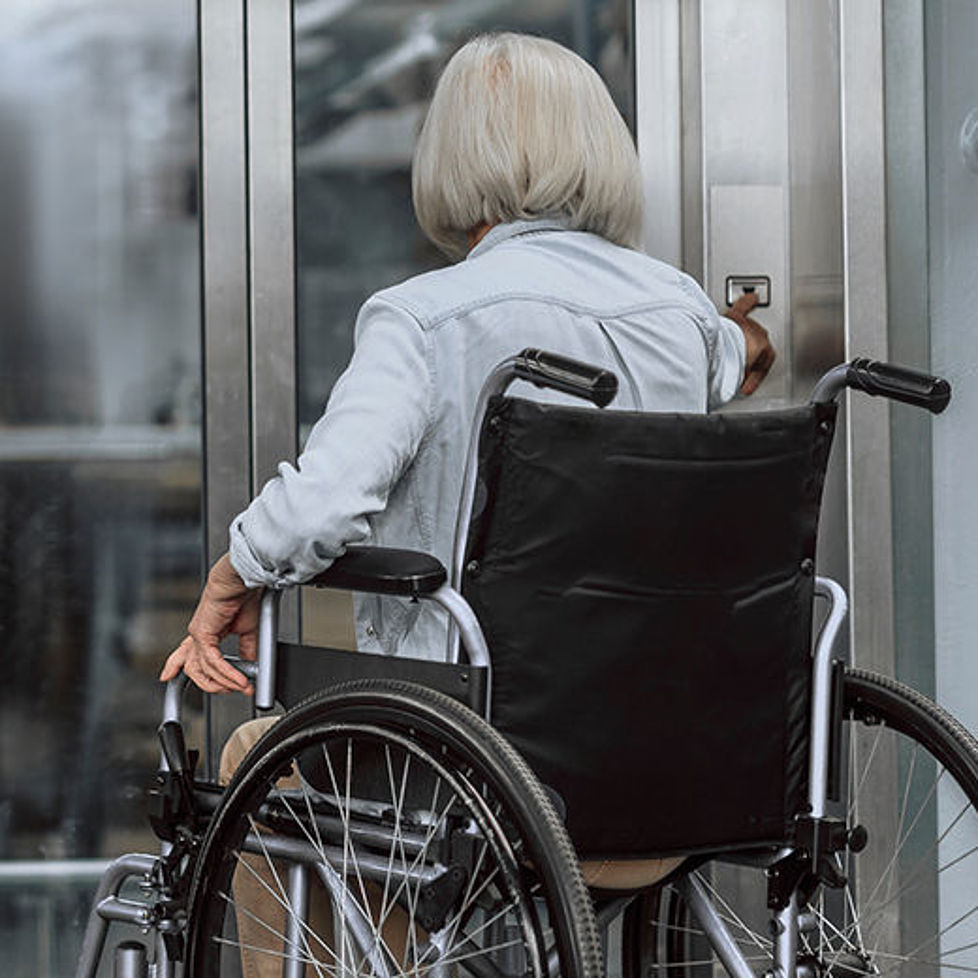 Eine ältere Frau in einem Rollstuhl befindet sich vor einer Fahrstuhltür und drückt auf den Ruf-Knopf.