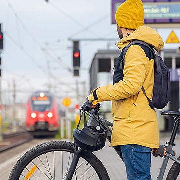 Ein Mann in einer gelben Regenjacke steht mit einem Fahrrad an an einem Bahnsteig, an dem ein roter Zug einfährt.