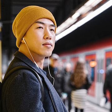 Ein Mann mizt gelber Mütze steht an einem Bahnsteig, ein roter Zug fährt ein.