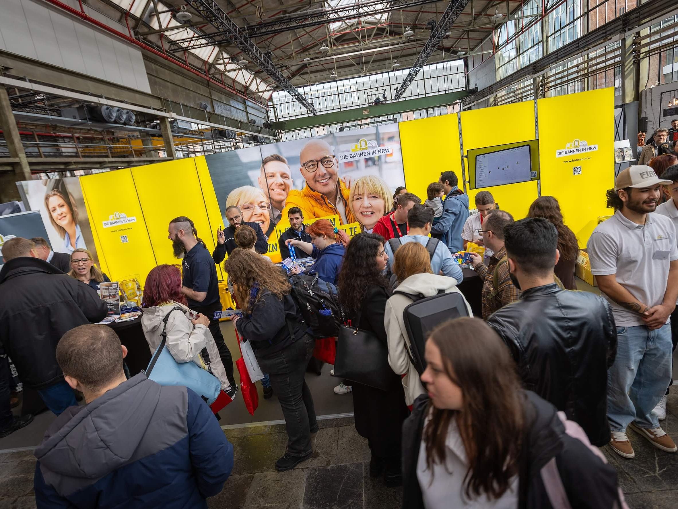 Das Bild zeigt Menschen in einer Messehalle. Im Hintergrund zu sehen ist die Wand eines Messestandes der Bahnen in NRW in gelb.