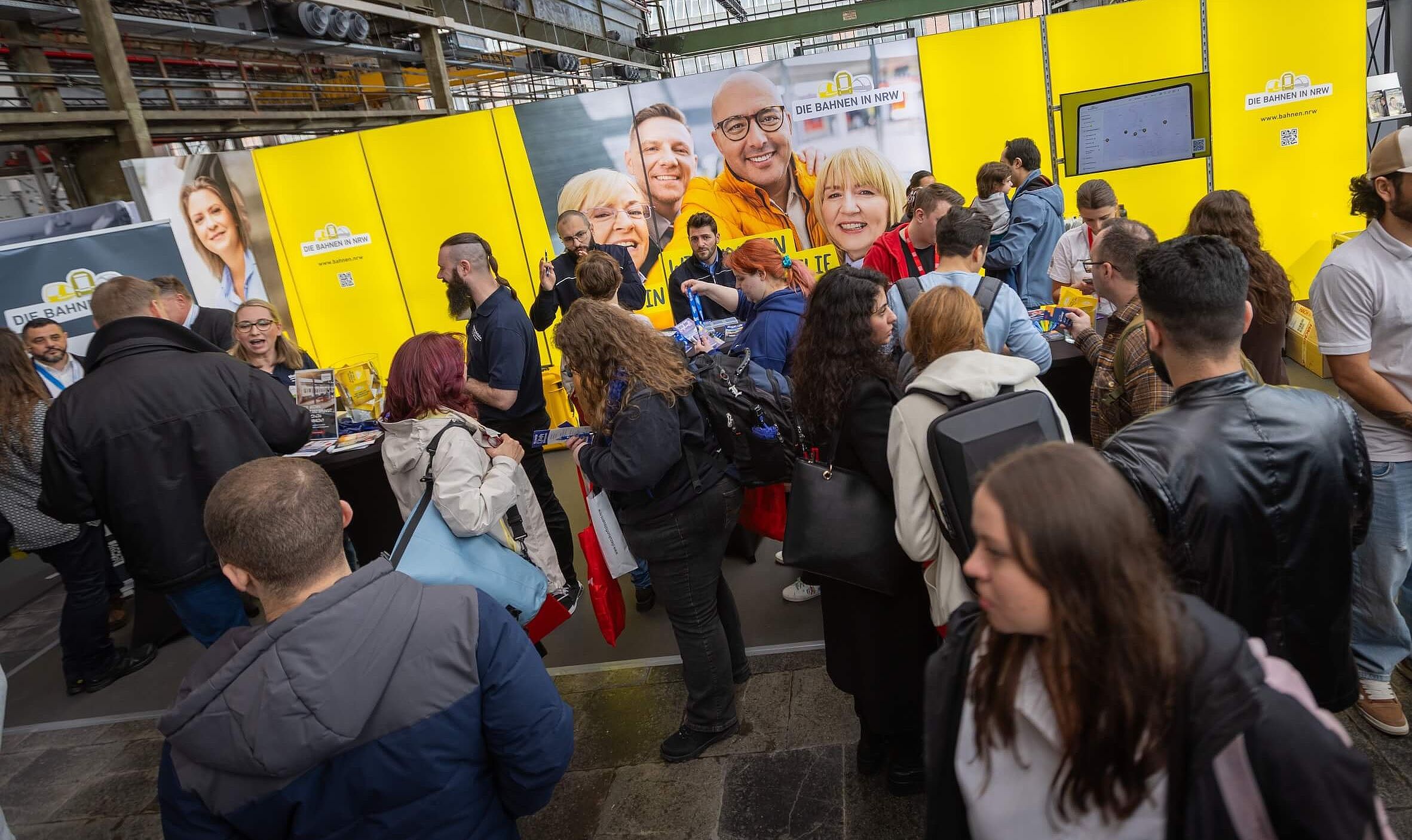 Das Bild zeigt Menschen in einer Messehalle. Im Hintergrund zu sehen ist die Wand eines Messestandes der Bahnen in NRW in gelb.